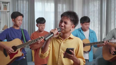 young asian boy singing and reading the lyrics on smartphone while his friends playing musical instruments at home