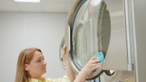 industrious woman in yellow shirt cleans washing machine door with blue soapy towel, carefully wiping glass surface of industrial laundry equipment to maintain cleanliness in modern laundromat