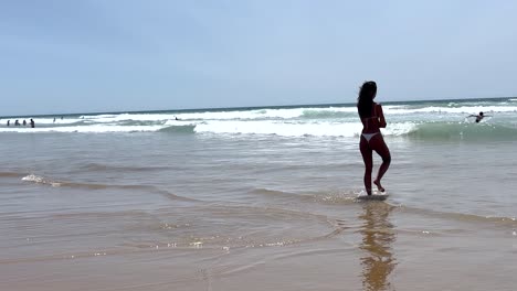 Una-Mujer-En-Bikini-Sumerge-Cautelosamente-Sus-Pies-En-El-Agua-Del-Océano,-Expresando-Dudas-Sobre-La-Temperatura-Del-Agua-Mientras-Disfruta-De-La-Experiencia-Junto-Al-Mar