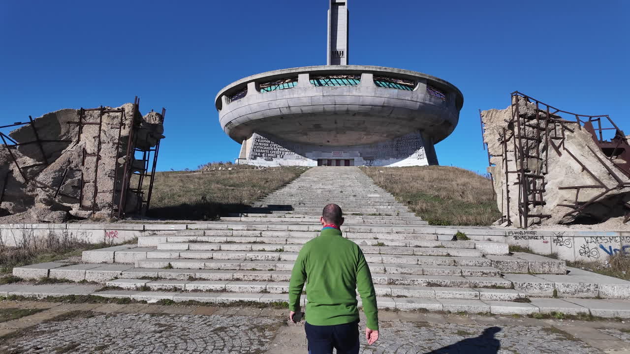 Tourist walking alone across windswept landscape toward massive abandoned Buzludzha communist era monument, iconic Bulgarian landmark standing desolate against dramatic mountain backdrop