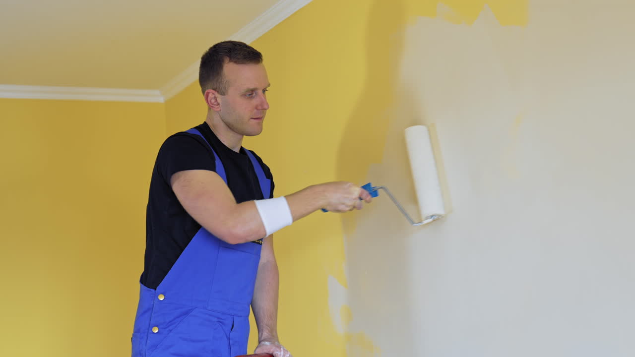 Professional painter paints the walls with roller brush. Worker man in blue overalls renovating and improving flat. Repair and decorating.