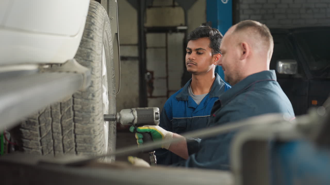 dos ingenieros automotores que trabajan en un taller mecánico, uno usando una herramienta neumática para quitar los pernos de las ruedas de un coche suspendido por un ascensor, mientras que el otro observa con una sonrisa, tocando su cabeza