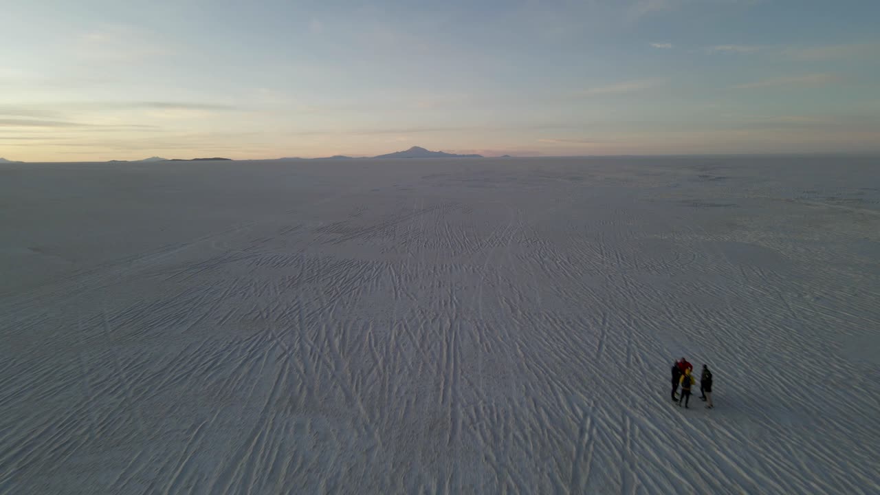 Sunset drone shot over mirror-like Uyuni salt flat with long reflections and soft orange tones as people admire the landscape