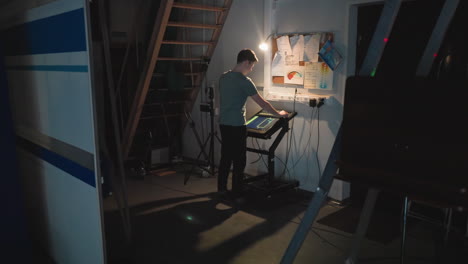 man stands under staircase using touchscreen control panel in dimly lit studio corner with tangled cords, technical gear, cork board of notes, and focused light source illuminating his workspace and tasks