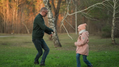 un hombre con una chaqueta verde y una niña pequeña con una jaqueta rosa, ambos con vaqueros azules, están alegremente saltando y jugando juntos al aire libre en un campo de hierba verde