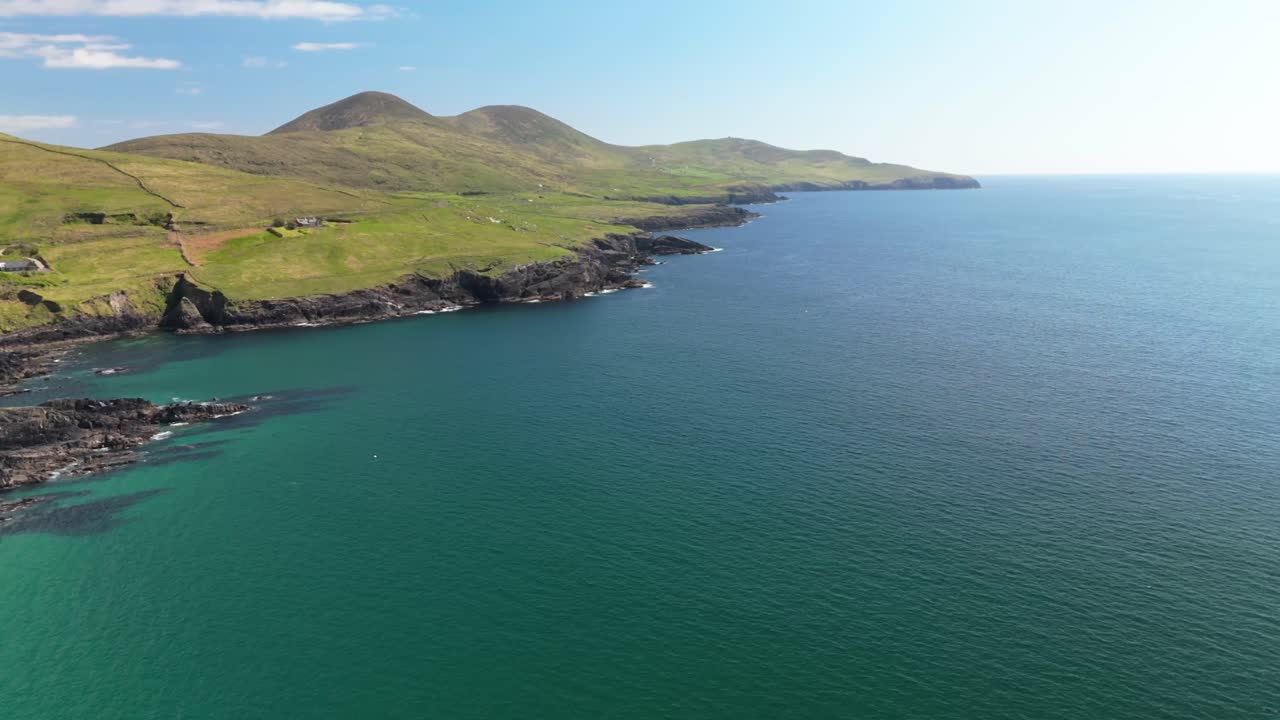 Drone flight backwards, with turn, along the Irish coastline reveals rugged cliffs, rolling green hills, and endless blue ocean.