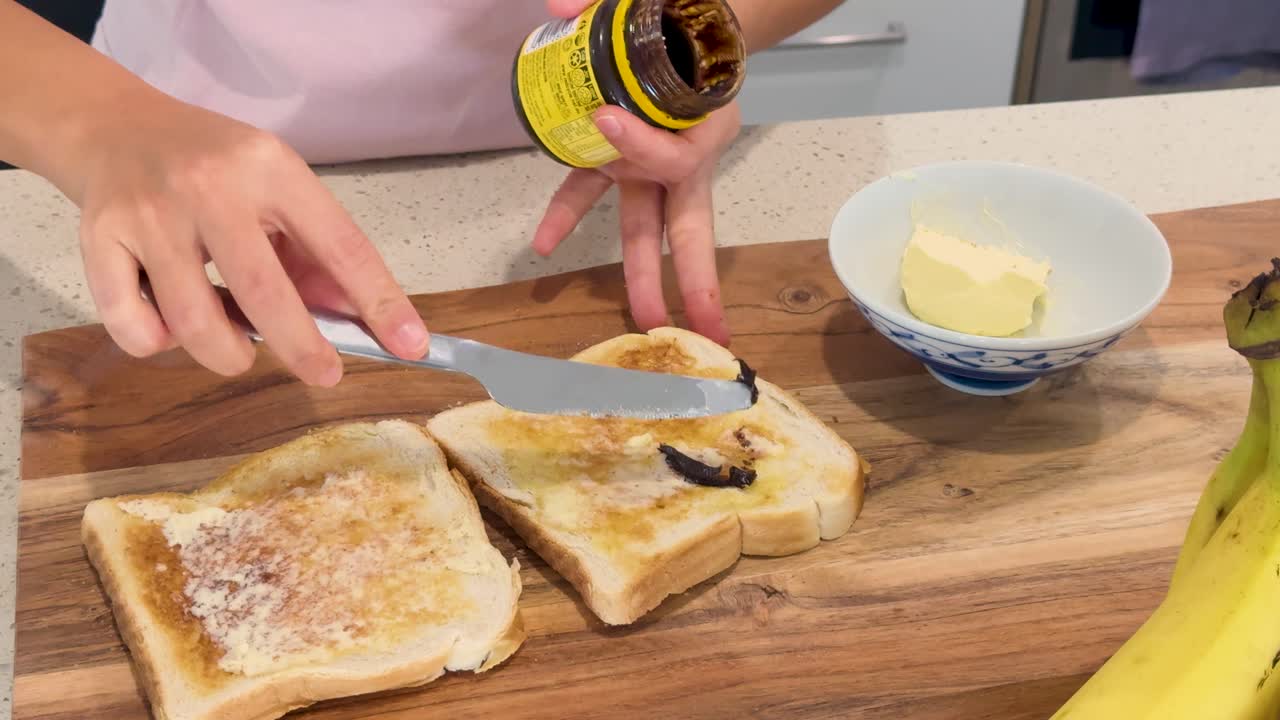 A person spreads dark yeast extract onto buttered toast slices on a wooden board in a brightly lit modern kitchen, using a butter knife
