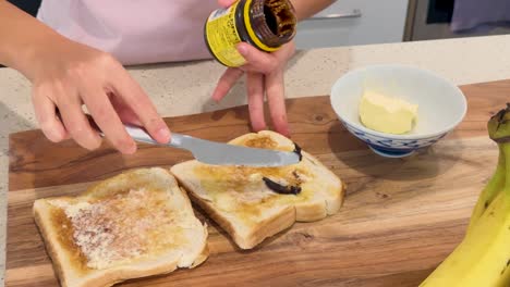 a person spreads dark yeast extract onto buttered toast slices on a wooden board in a brightly lit modern kitchen, using a butter knife