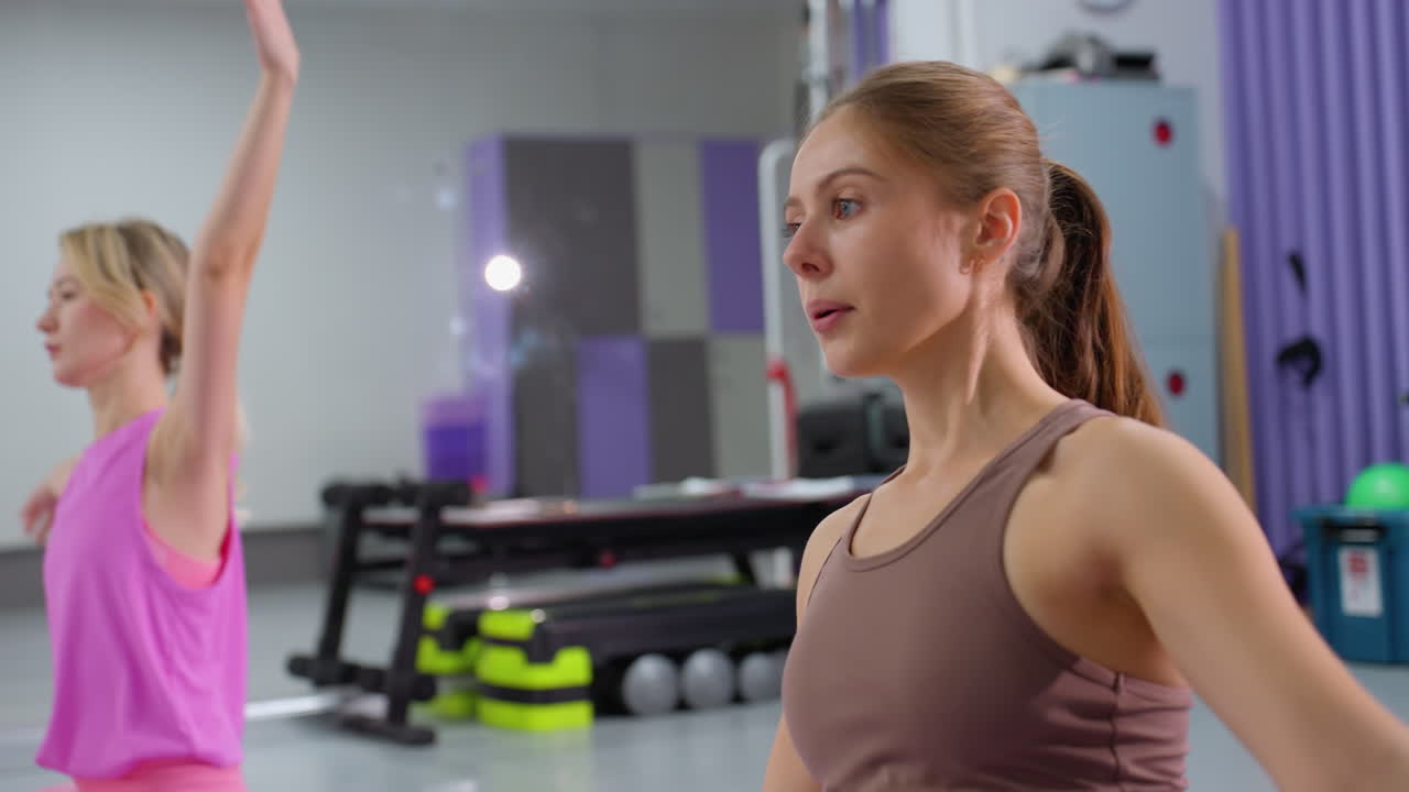 잘 장착 된 체육관에서 갈비 스트레칭 운동을 수행하는 매트에 앉아있는 여성, 배경에는 저항 밴드와 무게를 포함한 다양한 운동 장비가 있습니다.