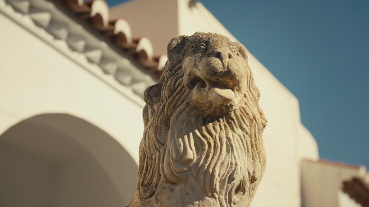 Scultura dettagliata di un leone in pietra illuminata da una calda luce solare, inquadrata da un edificio bianco in stile spagnolo e da un cielo blu in un'immagine drammatica dal basso