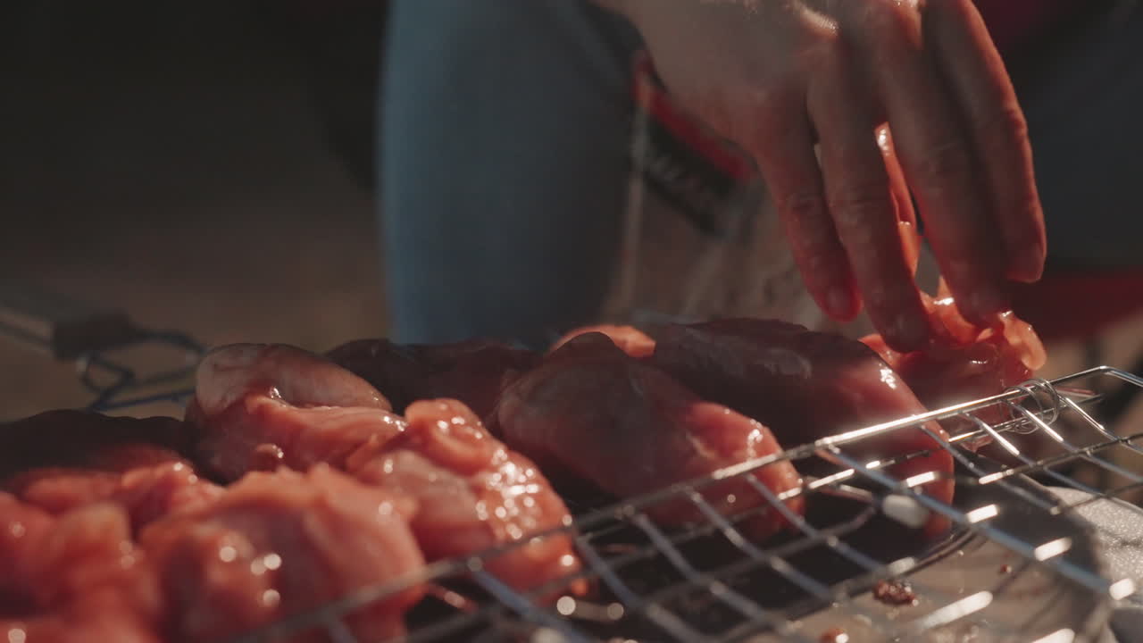 Tangan memarinasi ayam di panggangan kemping saat senja, close-up penuangan dari kantong plastik ke daging, gerakan membumbui yang terasa, panggangan logam mendesis, bara arang menyala, adegan memasak luar ruangan yang aktif