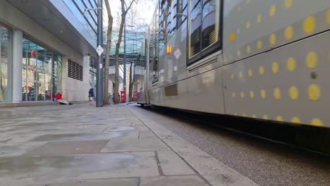 a yellow metrolink tram travelling down a street and under a glass footbridge with people passing by in manchester city centre