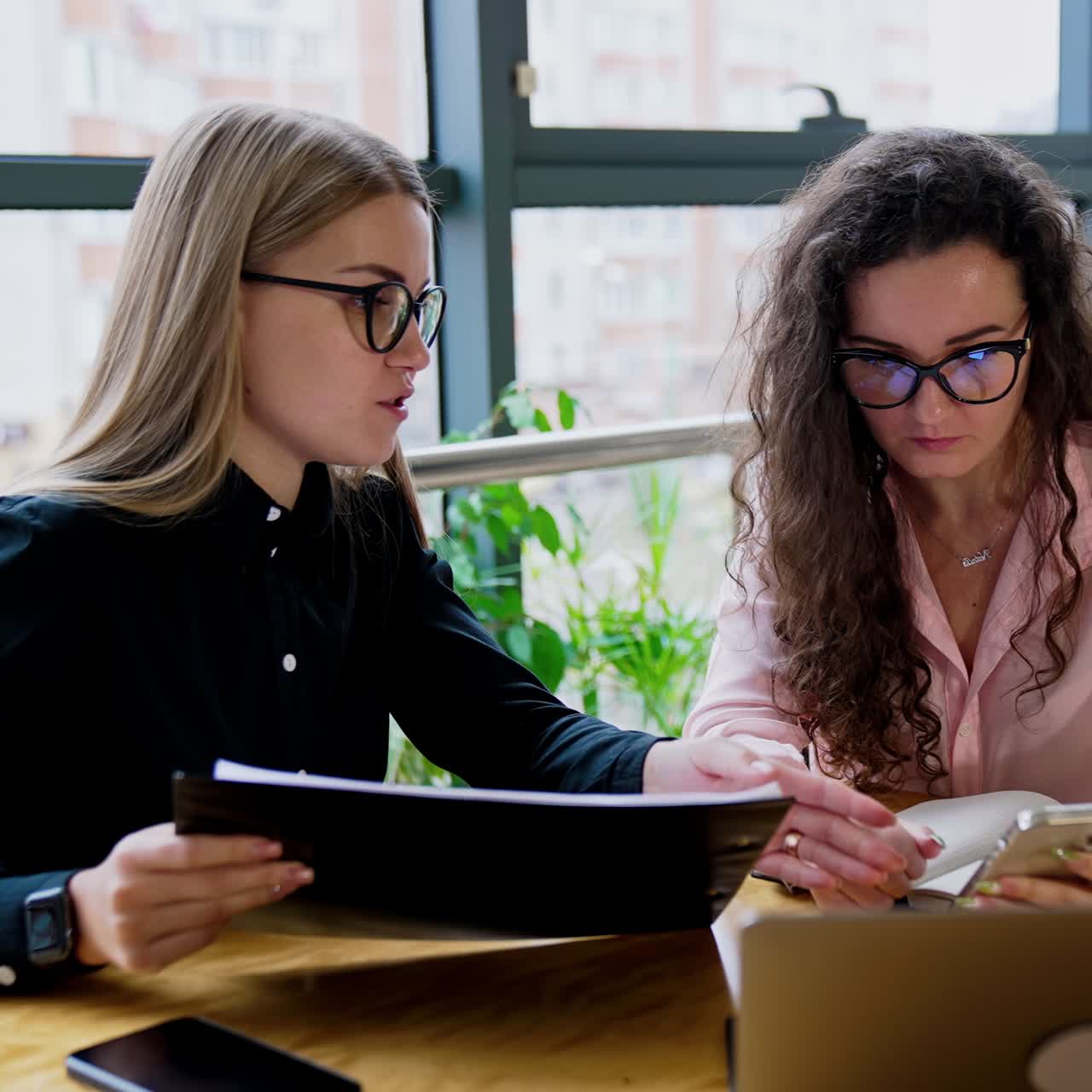 Young female colleagues talking about working issues sitting at the table. Ladies look at phone and have conversation