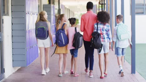 Diverse-female-teacher-and-happy-schoolchildren-walking-in-school