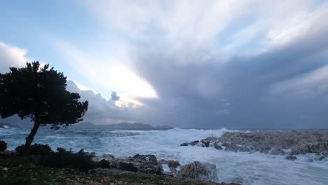 Deep-blue-waves-crash-on-rocky-shore-with-a-single-tree-and-dramatic-sky