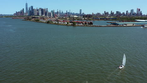 Una-Toma-Aérea-De-Un-Pequeño-Velero-Blanco,-Mientras-Pasa-Flotando,-El-Dron-Vuela-Arriba,-Con-El-Bajo-Manhattan-Al-Fondo-En-Un-Día-Soleado