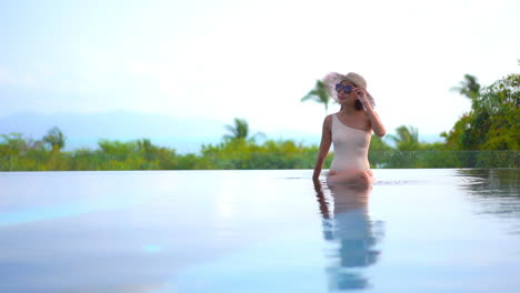 A-young-woman-sitting-in-the-shallow-end-of-an-infinity-edge-pool-reaches-up-to-adjust-her-sunglasses