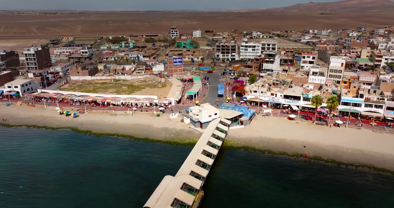 Peru'nun Paracas şehrinde kıyı kasabası binaları ve plajın yakınında okyanus suyuna uzanan uzun iskele. Havadan drone çekimi.