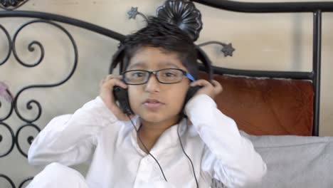 cute little indian asian caucasian boy with spectacles listening music on headphone dancing nodding head sitting in living room bedroom looking at camera front view