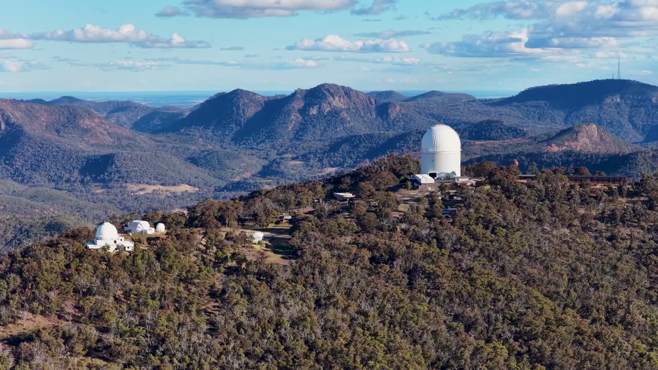 Drone camera glides over Siding Spring Observatory atop forested hills, revealing telescopes and rugged Warrumbungle mountain landscape under clear afternoon light