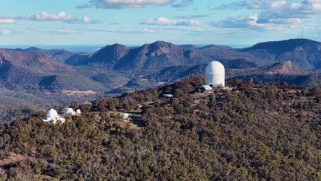 drone camera glides over siding spring observatory atop forested hills, revealing telescopes and rugged warrumbungle mountain landscape under clear afternoon light