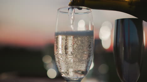 close up of wine pouring into glass held by hand with blurred person in background and shimmering bokeh lights under sunset sky capturing elegant pour motion and ambient evening mood