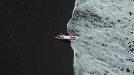 blond woman in white swimsuit lying on a dark beach while waves rooling around her