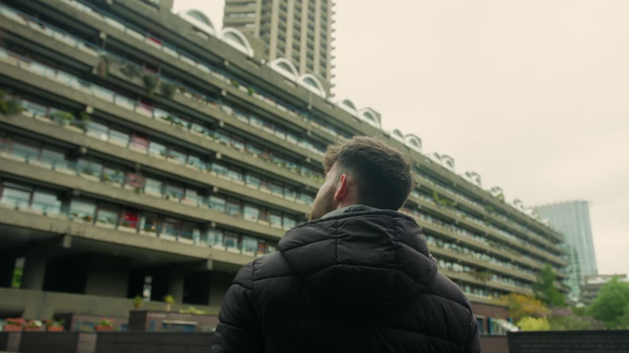 Rearview of man in black puffer jacket staring up below massive concrete walls in Barbican with heavy shadow and urban Brutalist style