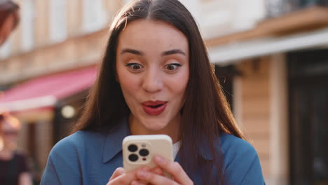 mujer asombrada usa teléfono inteligente celebrando ganar buen mensaje noticias lotería premio mayor victoria en la calle de la ciudad