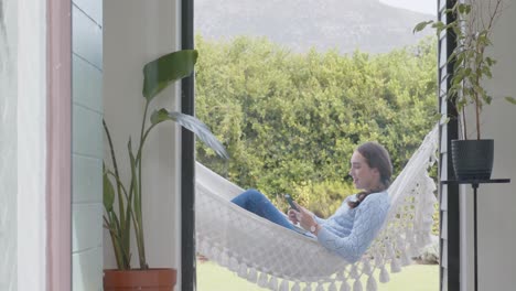 mujer caucásica feliz acostada en una hamaca y usando un teléfono inteligente en el jardín en cámara lenta