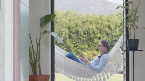 happy caucasian woman lying in hammock and using smartphone in garden in slow motion