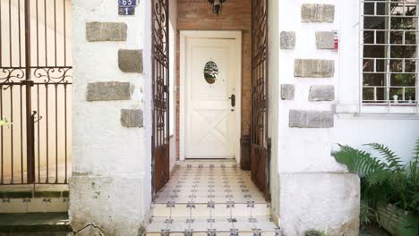 a white door with a small round window stands open in a charming entry framed by wrought iron gates, stone-accented walls, patterned floor tiles, and a nearby window with metal bars.