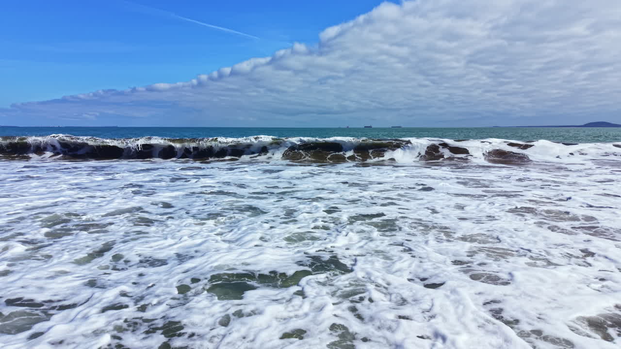 Vista di onde oceaniche che si infrangono su una spiaggia sabbiosa sotto un cielo blu con nuvole a mezzogiorno vicino alla zona costiera