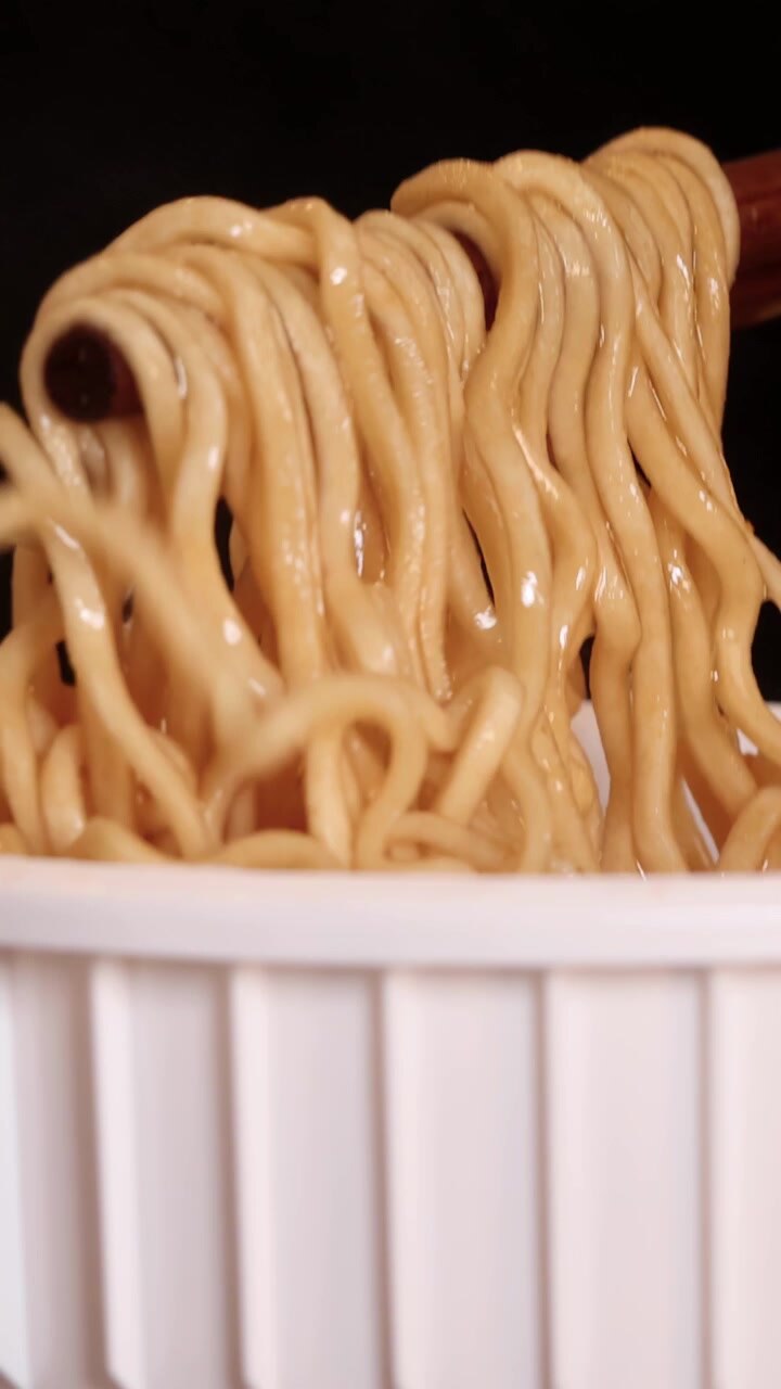 Bacchette sollevano noodles istantanei fumanti da una tazza bianca, primo piano con illuminazione drammatica.