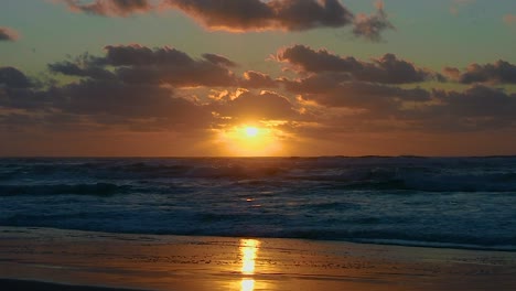 Dramatic-view-across-a-deserted-beach-with-the-sky-reflected-in-the-shoreline-and-a-stunning-golden-sunrise-or-sunset-with-the-low-sun-sparkling-off-the-rolling-and-foaming-surf