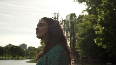 Attractive-young-Italian-woman-with-headphones-listening-to-music-at-the-park-with-a-sunset-look-around-smile,-dancing,-enjoying-life