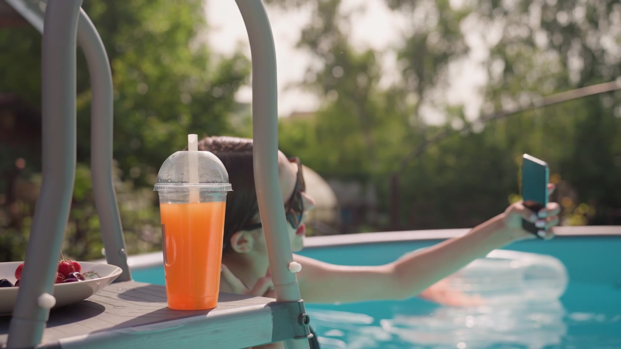 Young woman relaxes in pool capturing moments with her smartphone while enjoying orange drink and fresh fruit nearby. scene features bright outdoor lighting greenery background clear blue