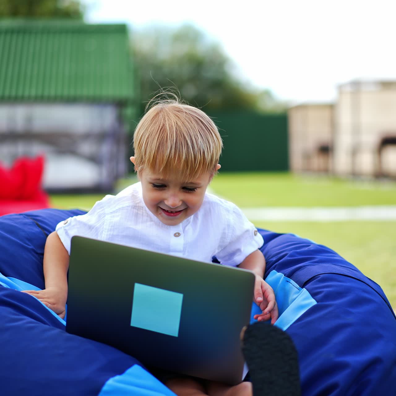 Nice little blond boy sitting comfortably in bean bag chair with laptop on his knees. Cute child spending time outdoors in summer. Blurred backdrop