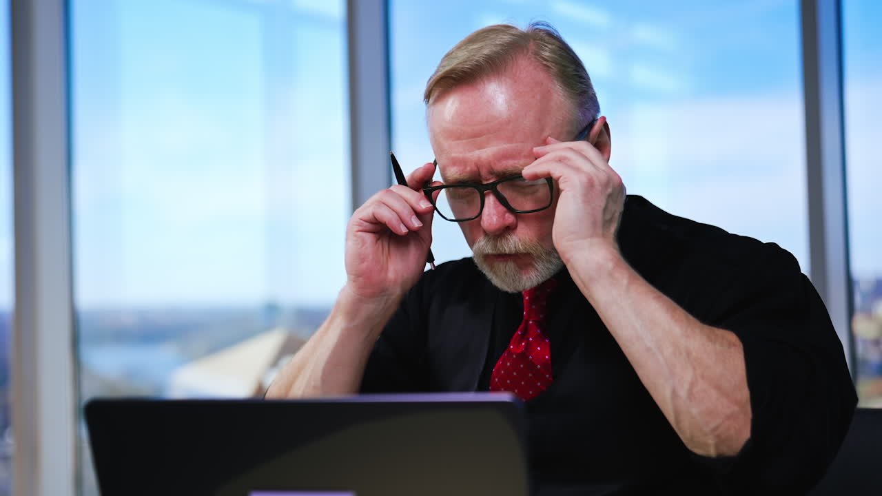 Old Caucasian businessman looks aside focused. Man puts on glasses and turns to his laptop. Close up.