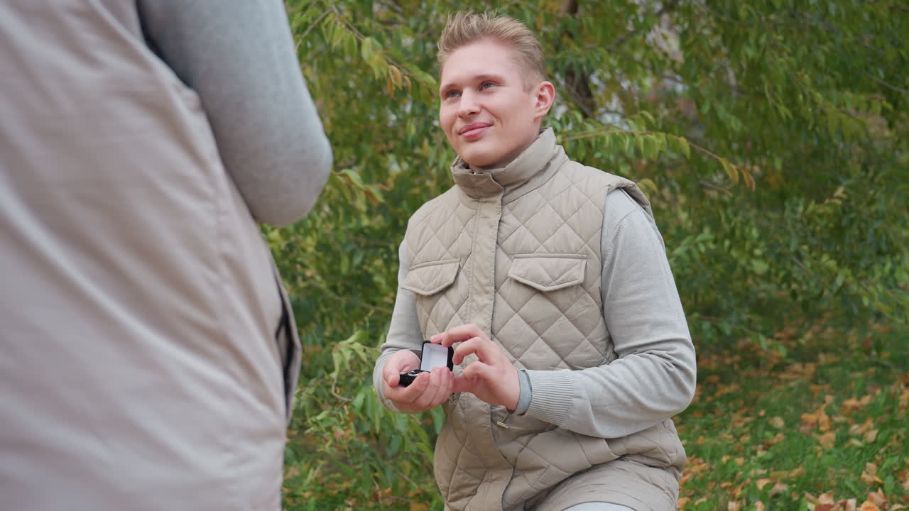 Heartfelt moment as man kneels to propose to his woman holding ring box with love and emotion in peaceful outdoor setting surrounded by greenery while both wear matching cozy outfits