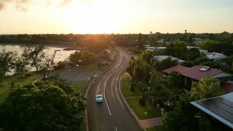 drone aerial of blue teal retro car down long winding road towards sunset