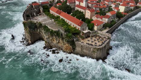 aerial fly iconic city on the sea cliff in budva montenegro with waves crushing around traditional architecture with red tile roofs, houses