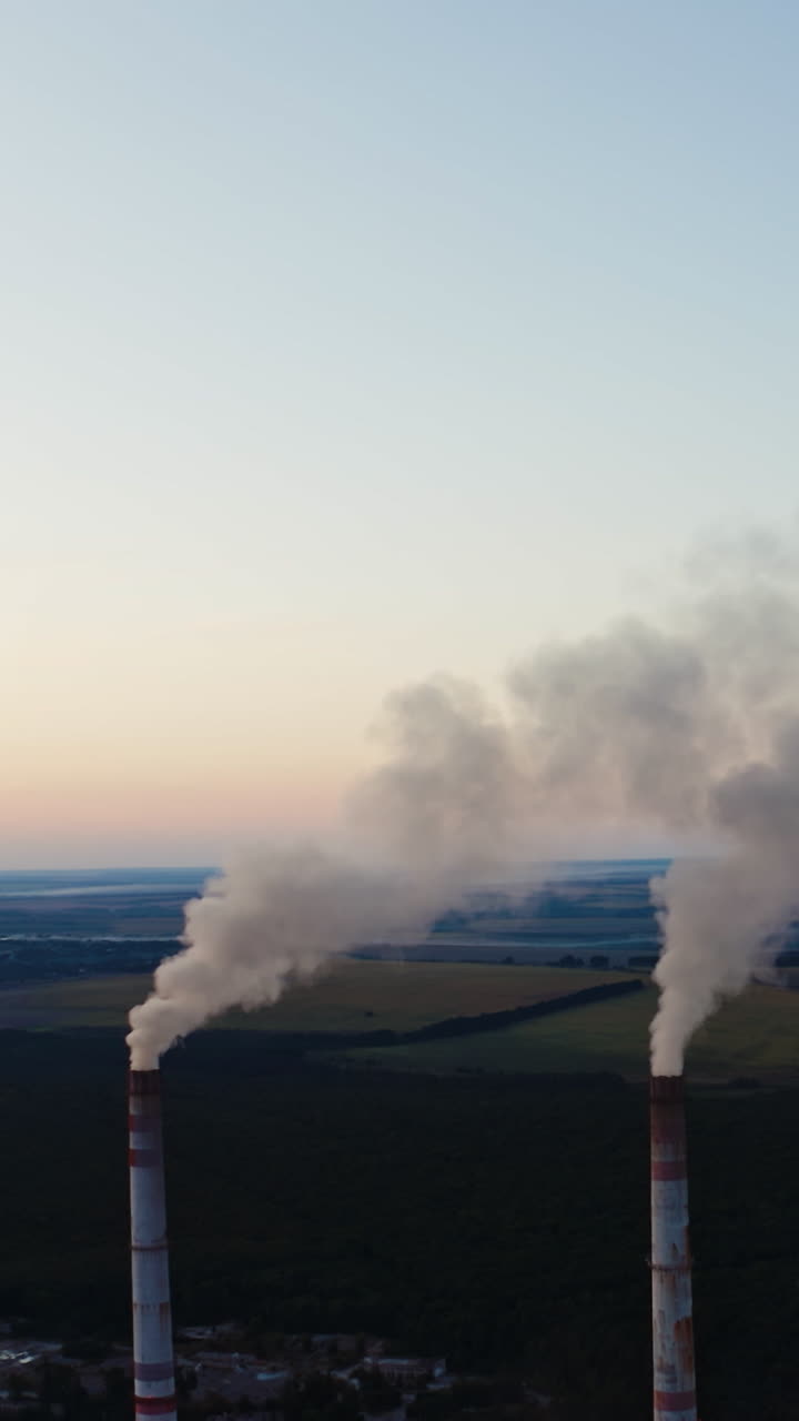 White smoke goes from pipes into the air. Industrial factory produce dirty emissions among the beautiful natural fields in the evening. Aerial view. Vertical video