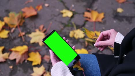 a woman's hand holds a mobile phone with a green screen while sitting in the park on a bench