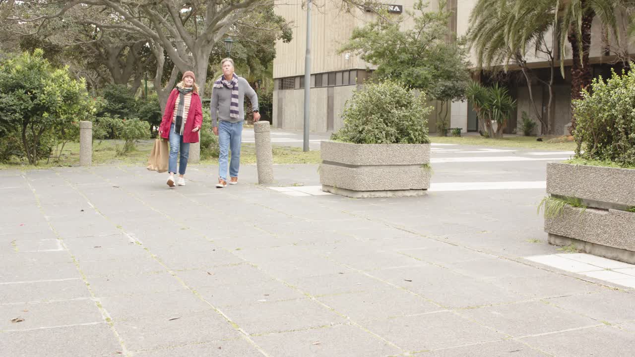 Coppia anziana che entra da sinistra e attraversa la piazza portando una borsa color cuoio e passando accanto ai dissuasori.