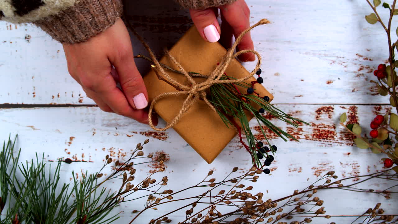 Hands tying a rustic gift with natural twine, decorated with pine sprigs and berries. Perfect for showcasing holiday gift-wrapping, DIY, and nature-inspired seasonal aesthetics. Flat Lay top view.