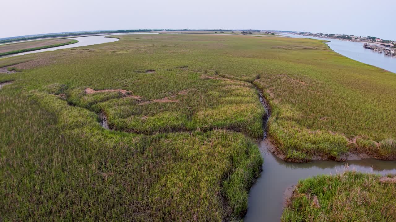 Drone footage captures green marshlands with winding waterways and tidal creeks near Charleston, South Carolina. The aerial view shows salt marsh vegetation and natural patterns under a pastel sky