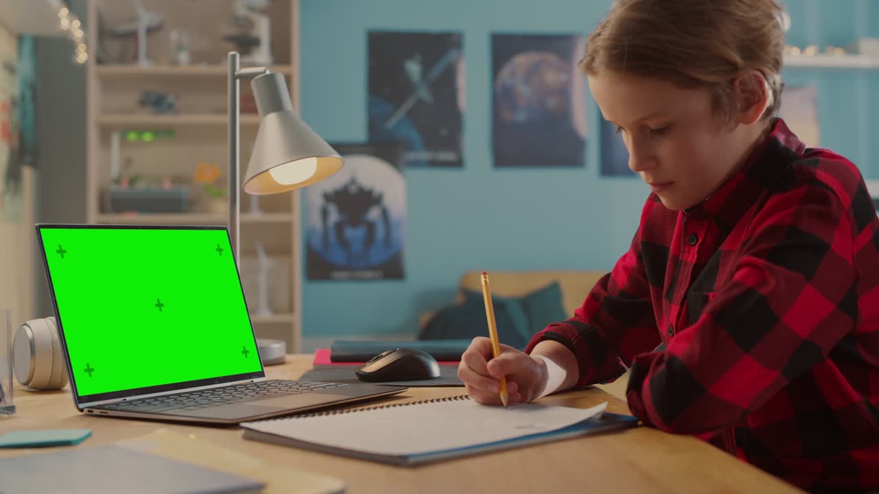 un joven inteligente con auriculares asistiendo a una clase en línea en una computadora portátil con pantalla verde en casa. un adolescente feliz navegando por la investigación educativa en línea, escribiendo en un cuaderno, estudiando la tarea escolar.