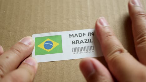 Manos-Aplicando-La-Etiqueta-De-La-Bandera-Hecha-En-Brasil-En-Una-Caja-De-Envío-Con-Un-Código-De-Barras-De-Primera-Calidad-Del-Producto