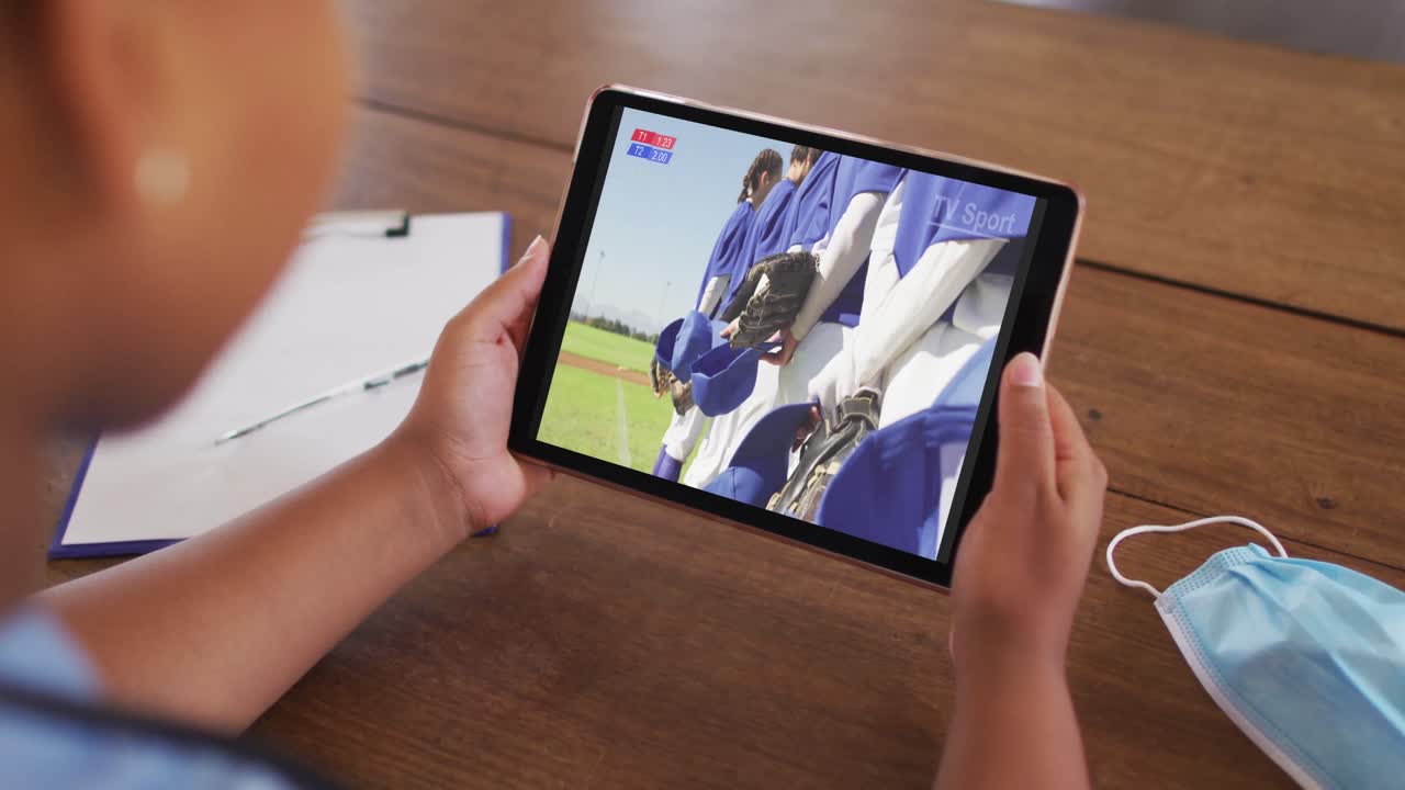 태블릿에서 야구 경기를 시청하는 얼굴 마스크와 함께 테이블에 앉아있는 여성의 복합